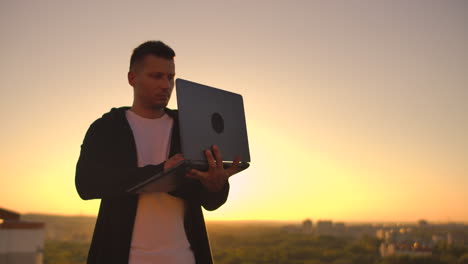 A-man-with-a-laptop-at-sunset-stands-on-the-roof-of-the-building-and-writes-a-code-on-the-keyboard.-Hacker-with-laptop.